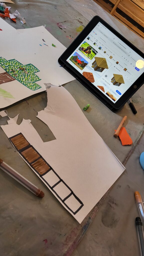 Das Bild zeigt eine Situation mit Bastelmaterialien wie Schere, Stifte, Papier und ein Tablett, auf dem Vorlagen für Minecraft-Elemente zu sehen sind. Diese werden gerade nachgemalt und ausgeschnitten.